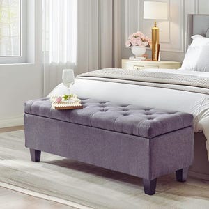 May include: A grey upholstered storage bench with button detailing, set on dark wooden legs. The bench is in a bedroom, with a bed and bedside table in the background. A glass and a tray are on top.