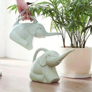 以下が含まれることがあります： 2つの薄緑色のゾウの形をしたじょうろ。1つは手に持たれ、もう1つは木製の表面に置かれています。じょうろには鼻の注ぎ口と上部にハンドルが付いています。背景には鉢植えの植物があります。