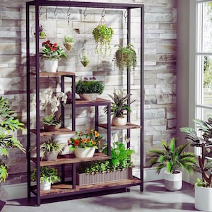 Op de afbeelding: Een plantenstandaard van zwart metaal en hout met meerdere planken en ophanghaken. De planken tonen diverse potplanten, waaronder bloeiende planten, varens en vetplanten. De standaard staat tegen een stenen muur, wat een natuurlijke uitstraling geeft.