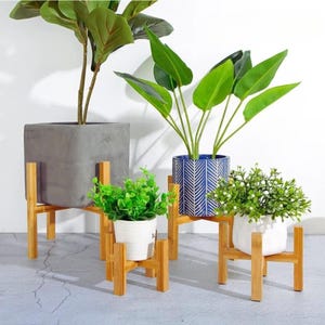 May include: Four wooden plant stands of varying heights, each holding a different potted plant. The plants include a large-leafed green plant, a small green plant, and a plant with white flowers. The stands are made of light-coloured wood.