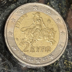 Puede incluir: Una moneda de dos euros con un centro dorado y un anillo exterior plateado. El centro dorado presenta a una mujer montando un toro, con el texto "2 EURO" y el año "2002". La moneda está rodeada de estrellas.
