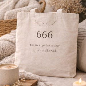 Może przedstawiać: Beżowa torba typu tote z numerem 666 pośrodku, z motywem skrzydeł nad nim i tekstem "You are in perfect balance. Trust that all is well." Torba ma dwa uchwyty.