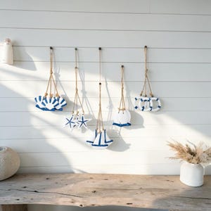 Può includere: Decorazioni a tema costiero appese in bianco e blu. La collezione comprende forme di conchiglie, stelle marine, barche e salvagenti, sospese con spago naturale contro un muro bianco. Un tavolo in legno rustico e un vaso con fiori secchi sono in primo piano.