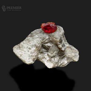 May include: A faceted, deep red gemstone rests atop a rough, gray mineral formation. The gemstone is the focal point, catching the light and displaying a vibrant color. The mineral has a layered texture.