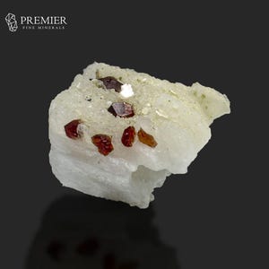 Puede incluir: Primer plano de un espécimen mineral con pequeños cristales de color marrón rojizo incrustados en una matriz de color claro. Los cristales tienen un aspecto brillante y la matriz tiene una superficie rugosa y texturizada. El mineral se encuentra sobre un fondo oscuro.