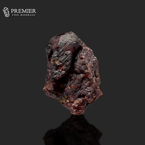 Könnte beinhalten: Ein raues, dunkelrotes Mineralexemplar mit kristalliner Struktur. Das Logo von Premier Fine Minerals befindet sich in der oberen linken Ecke. Das Mineral hat eine reflektierende Oberfläche vor dunklem Hintergrund.