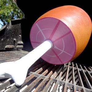 Pode incluir: Um grande acessório de presunto de madeira pintada, repousando sobre uma grelha. O presunto é laranja e rosa, com um osso branco. A grelha é preta e enferrujada, com um fundo natural.