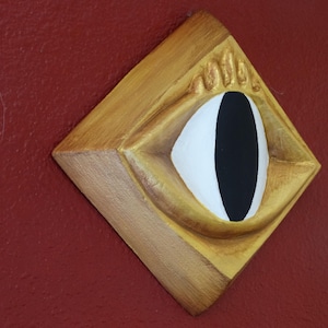 Peut inclure: Une œuvre d'art murale en bois en forme de losange représentant un œil stylisé. L'œil a un iris blanc et une pupille noire, encadré dans un cadre doré. Le fond est rouge foncé.