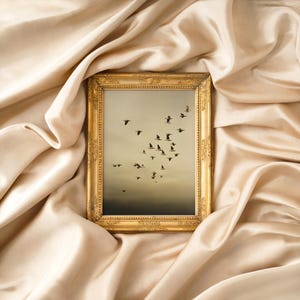Puede incluir: Una obra de arte enmarcada que representa una bandada de pájaros en vuelo contra un cielo apagado. El marco dorado tiene detalles ornamentales. La obra se exhibe sobre un fondo de tela drapeada de color crema.