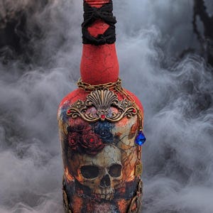 May include: A decorative glass bottle with a red crackle finish, featuring a prominent skull and rose graphic. The bottle is adorned with a black fabric neck wrap, a faceted black stopper, ornate bronze-toned metal filigree, and a hanging chain.