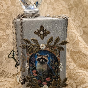 May include: A rectangular silver-toned decorative bottle topped with a large faceted clear crystal stopper. The front features a central framed illustration of a raccoon surrounded by flowers, accented with bronze-colored metal leaves, rhinestones, and a dangling chain with small metal keys.
