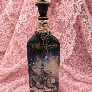 May include: A decorative black bottle featuring a dark finish with gold-toned filigree accents at the top and bottom. The center displays a graphic of two lace-up boots, adorned with a dangling bronze chain, a purple crystal, and small clear rhinestones.