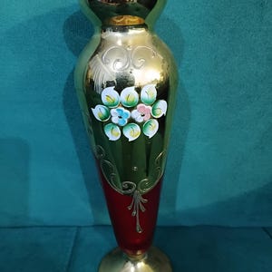 May include: A decorative vase with a gold-colored top and base, and a red center. The vase features hand-painted floral designs in white, blue, and pink. The vase is approximately 10 inches tall.