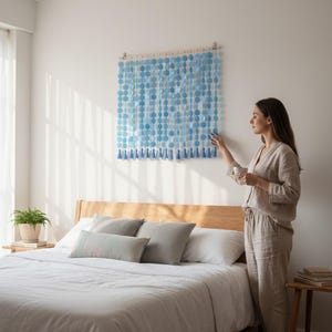 Könnte beinhalten: Ein hellblaues Wandbehang mit kreisförmigen Aussparungen und Quasten, über einem Bett mit weißer Bettwäsche und grauen Kissen. Der Wandbehang besteht aus mehreren Strängen blauer Kreise. Eine Frau steht neben dem Bett.