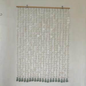 Peut inclure: Une tenture murale décorative composée de ronds blancs en forme de coquillages suspendus à une tringle en bois. Le bas de la tenture est orné de petits pompons vert clair. La tenture est conçue pour ajouter une esthétique naturelle et côtière.