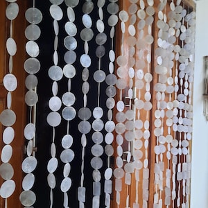 May include: A beaded door curtain made of white, iridescent, circular and rectangular shell pieces. The curtain hangs in front of a wooden door frame. The beads are strung together with white string, creating a decorative and natural look.