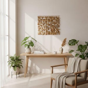 Könnte beinhalten: Ein quadratischer Wandbehang, der aus zahlreichen kleinen, runden, hellbraunen Holzscheiben besteht, die an vertikalen Schnüren an einer horizontalen Stange hängen und eine texturierte, geschichtete Optik an einer schlichten Wand erzeugen.