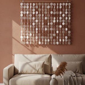 May include: A decorative wall hanging made of numerous, small, round, white, shell-like discs suspended from a wooden rod. The wall hanging is displayed against a peach-colored wall above a beige sofa. The room has a minimalist, bohemian aesthetic.