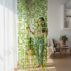 Pode incluir: Uma cortina de contas verdes e circulares está pendurada em frente a uma janela. A cortina é feita de muitas contas pequenas, redondas e verde claro. Uma pessoa está atrás da cortina. O quarto tem uma paleta de cores neutras.