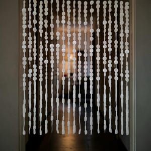 May include: A white beaded curtain hangs in a doorway, with strands of varying lengths. The beads are round and oval, creating a textured, semi-transparent barrier. The background shows a dimly lit hallway.