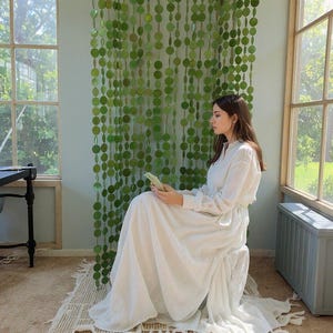 May include: A long-sleeved white maxi dress with a gathered waist and a full, flowing skirt. The garment is made of a lightweight, semi-sheer fabric and features a simple collar. It is positioned in front of a decorative green circular disc curtain.