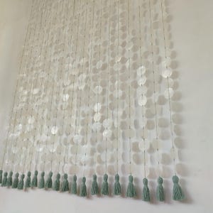 Op de afbeelding: Een hangend scheidingsgordijn bestaande uit meerdere verticale strengen van doorschijnende, ronde witte schelpschijfjes. Elke individuele streng eindigt in een zachte, saliegroene stoffen kwast, wat een gelaagd, gestructureerd en lineair visueel effect creëert.