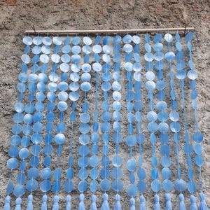 Puede incluir: Una cortina de cuentas azul claro con una barra de madera. La cortina está hecha de cuentas redondas y planas ensartadas en filas verticales. Cada fila termina con una borla. La cortina cuelga contra una pared gris texturizada.