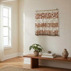 Peut inclure: Une tenture murale composée de multiples brins verticaux de disques circulaires translucides blancs et brun clair suspendus à une tige en bois horizontale. Le bas de chaque brin présente un petit pompon en tissu brun clair.