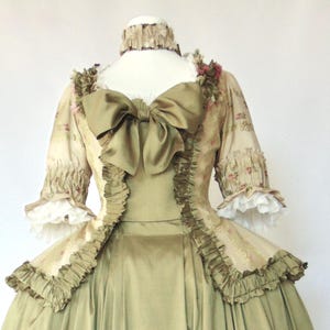 May include: A vintage-style olive green gown with a large bow at the back. The dress features a fitted bodice, ruffled trim, and a full skirt. The sleeves are short and adorned with lace cuffs. The gown is displayed on a mannequin.