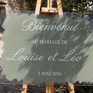May include: A rectangular clear acrylic wedding sign featuring a sage green painted brushstroke background. The white text reads Bienvenue AU MARIAGE DE Louise et Léo 2 MAI 2026. The sign is displayed on a wooden easel, showcasing a clean and modern design.