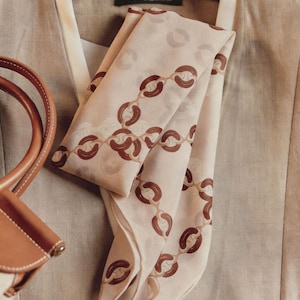 Puede incluir: Una bufanda beige con un estampado de cadena marrón está drapeada sobre una blazer beige. La bufanda está doblada, revelando el diseño de la cadena. Un bolso de mano de cuero marrón es visible en la esquina. La palabra "VOGUE" está impresa en una etiqueta.
