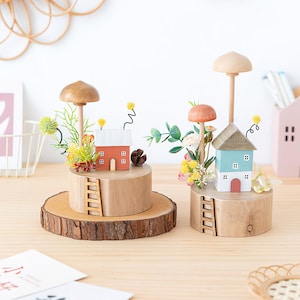 Pode incluir: Duas casas decorativas de madeira com tampas em forma de cogumelo. Uma casa é laranja, a outra azul. Ambas têm pequenas escadas, detalhes florais e assentam sobre bases de madeira. As bases estão sobre uma mesa de madeira.