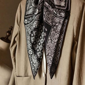 Puede incluir: Un pañuelo negro y blanco con estampado de cachemira drapeado sobre una blazer beige. El pañuelo presenta intrincados diseños florales y de lágrimas. La blazer tiene un corte clásico con cierre de botón y un bolsillo. El pañuelo añade un contraste audaz.