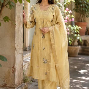 May include: A light yellow three-piece ensemble consisting of a long-sleeved kurta with a V-neckline, matching wide-leg trousers, and a sheer dupatta. The kurta features delicate floral embroidery in pink and green tones scattered across the fabric.