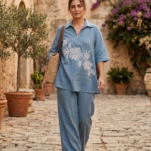 Könnte beinhalten: Hellblaues Leinen-Outfit, bestehend aus einem kurzärmeligen Oberteil mit floraler Stickerei und einer passenden Hose mit weitem Bein. Das Oberteil hat einen Reißverschlusskragen. Das Outfit wird mit einer Strohtasche und braunen Sandalen kombiniert. Im Hintergrund ist ein Steinweg zu sehen.