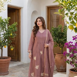 Pode incluir: Um vestido túnica malva de manga comprida com decote em V, pregas verticais no corpete e bordados florais creme na parte inferior. O conjunto inclui calças a condizer e um xaile transparente com rebordo dourado.