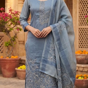 May include: A slate blue three-piece ensemble featuring a long-sleeved tunic with silver embroidery along the neckline and hem, matching straight-leg trousers, and a sheer, lightweight dupatta with a decorative border and floral embroidery.