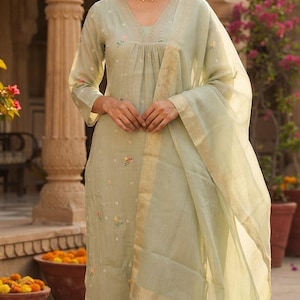 May include: A light sage green three-piece ensemble consisting of a V-neck kurta with delicate floral embroidery, matching straight-leg trousers, and a sheer dupatta featuring a gold metallic border and tassel details at the hem.