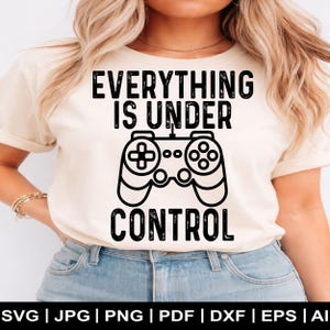 May include: A cream-colored t-shirt with the text "EVERYTHING IS UNDER CONTROL" in black, along with a graphic of a video game controller. The person is wearing blue jeans.