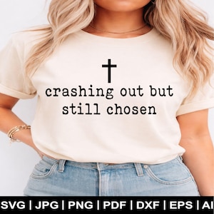 以下が含まれることがあります： 黒い十字架が上に配置された、黒い文字で「crashing out but still chosen」と書かれたクリーム色のTシャツ。シャツはブルージーンズと組み合わされています。画像の下部にはファイルタイプのオプションが表示されています。