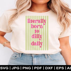 May include: Cream-colored t-shirt with the text "literally born to dilly dally" in pink, set against a vertical striped background of light green and white. The shirt is a casual style.