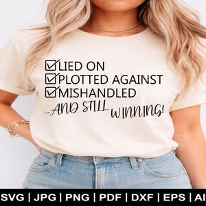 May include: A cream-colored t-shirt with the text "LIED ON, PLOTTED AGAINST, MISHANDLED...AND STILL WINNING!" in black. The shirt has a classic crew neck and short sleeves. The bottom of the image shows file type options.