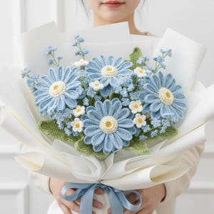May include: A bouquet of crocheted flowers featuring large blue daisies with yellow centers, small white flowers, and blue sprigs, wrapped in white paper and tied with a blue ribbon. The flowers have green leaves.