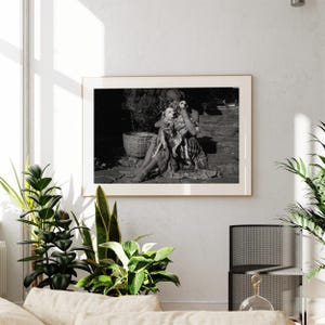a black and white photograph of a woman sitting on a bench, holding a dog in her lap. The photograph is framed and hung on a wall, with several potted plants placed around the room.