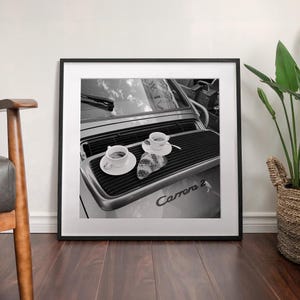 A framed black and white photograph of a car with two cups of coffee on the hood, placed on a wooden floor next to a woven basket.