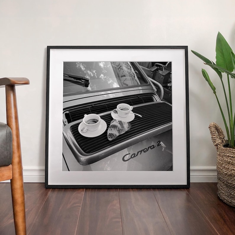 A framed black and white photograph of a car with two cups of coffee on the hood, placed on a wooden floor next to a woven basket.