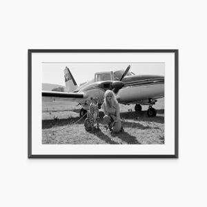 A black and white photograph of a woman kneeling next to a cheetah in front of an airplane.