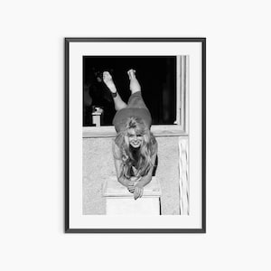 A black and white photograph of a woman posing with her legs crossed and a smile on her face, leaning against a window frame.