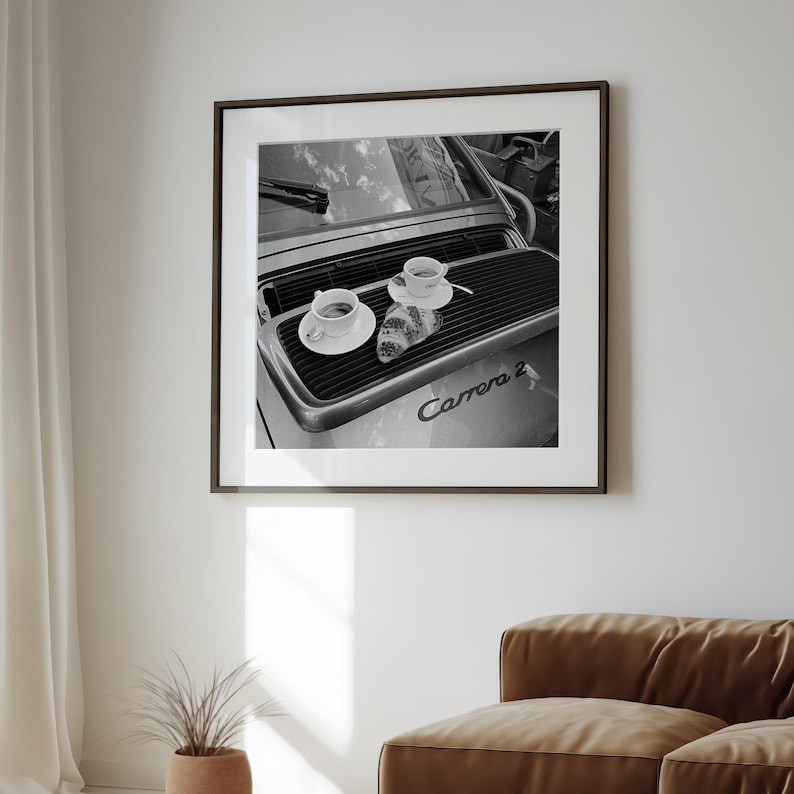 A framed black and white photograph of a car with two cups of coffee on the hood, hanging on a wall in a living room.