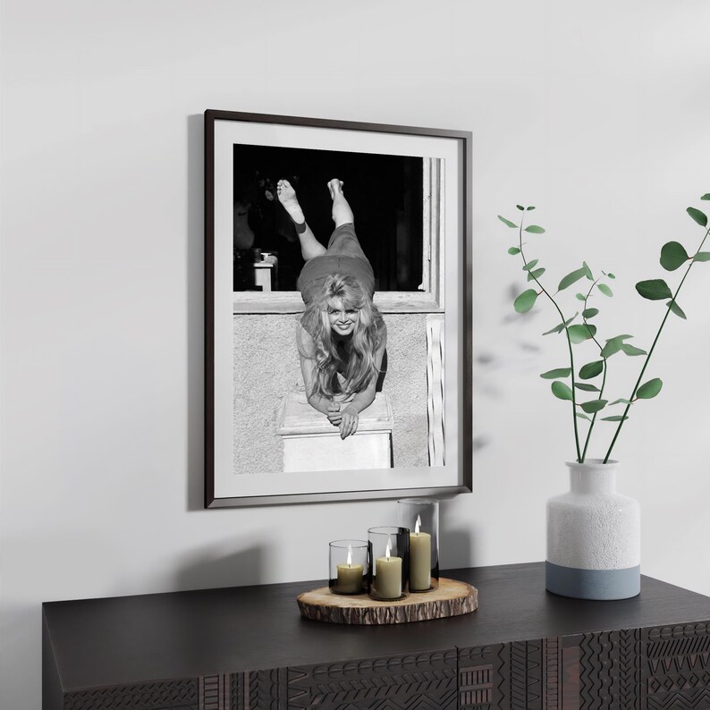 a framed black and white photograph of a woman leaning on a window sill, with a vase of greenery and candles on a wooden surface below the frame.
